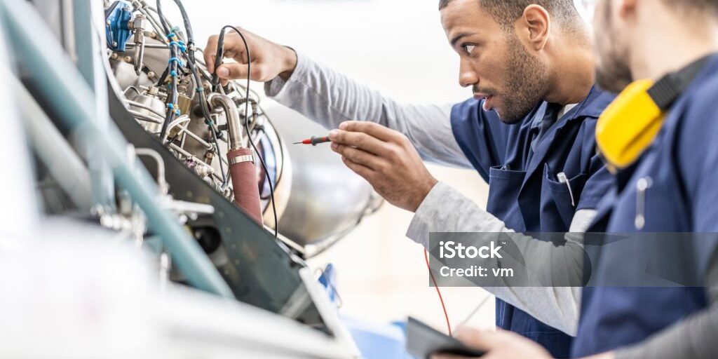 aerospace-is-1403809667-1024x1024 Aircraft mechanics examining helicopter engine with multimeter, using multimeter, side view. Engineers working in hangar together, medium shot